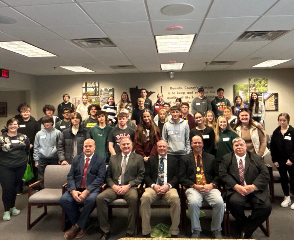 A large group of students posing with five county commissioners in a board room.