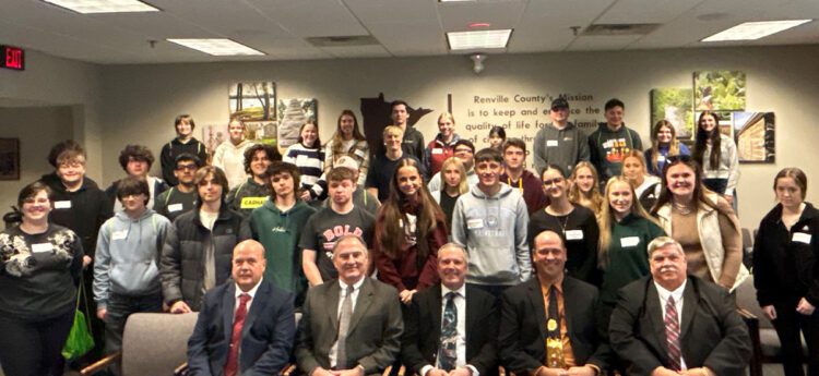 A large group of students posing with five county commissioners in a board room.